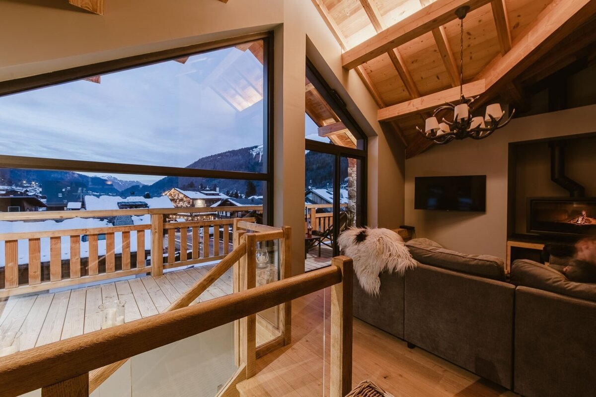 A warm, modern chalet interior with exposed wood beams, a fireplace, and large windows revealing a snowy mountain village nestled among hills.