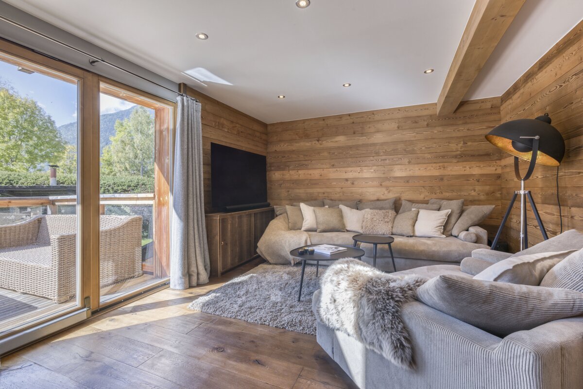 A cozy, wood-paneled living room with plush sofas, a TV, and large glass doors leading to a balcony with mountain and tree views. Warm wooden floors complete the look.