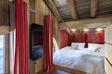 A cozy, rustic bedroom with wood-paneled walls, exposed beams, a red patterned headboard, a white bed with teddy bears, and red striped curtains.