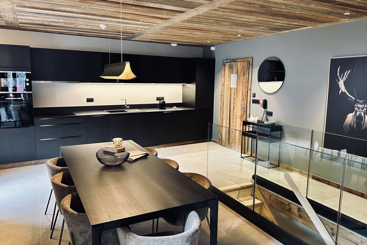 A stylish modern kitchen with black cabinets and dining table, featuring a rustic wooden ceiling and a glass-railed staircase descending.