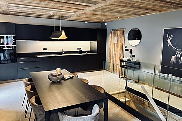 A stylish modern kitchen with black cabinets and dining table, featuring a rustic wooden ceiling and a glass-railed staircase descending.