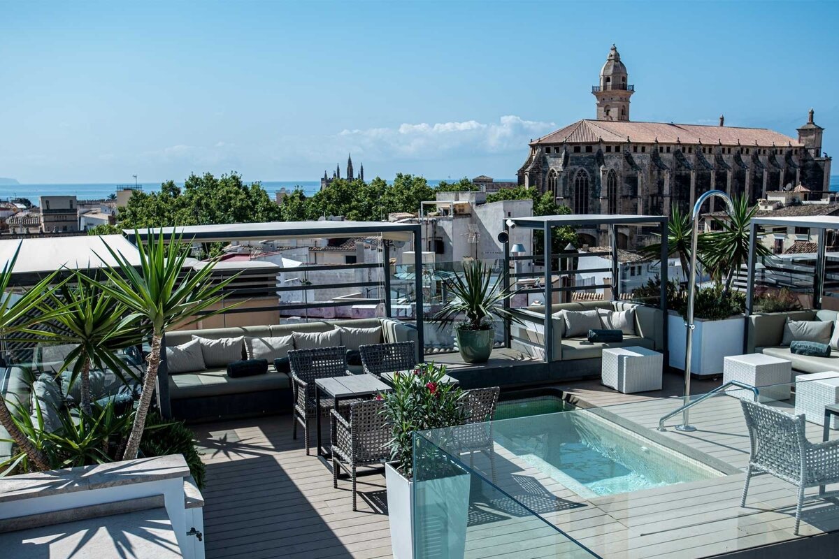 Luxurious rooftop terrace with a jacuzzi, lounge, and dining, overlooking a historic city with a church and the sea.