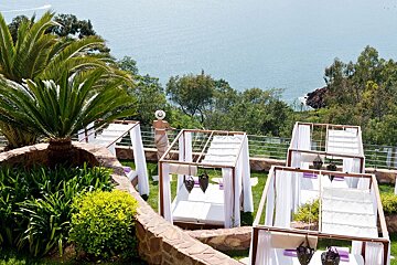 A woman in a white hat stands in a lush green garden overlooking the ocean