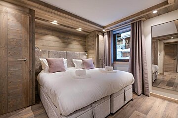 A cozy, wood-paneled bedroom with a large bed, pink pillows, window overlooking a snowy village, and a full-length mirror.