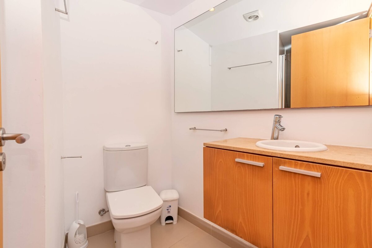 A clean, modern bathroom with a white toilet, wooden vanity, sink, and large mirror, set against white walls and light-colored floors.