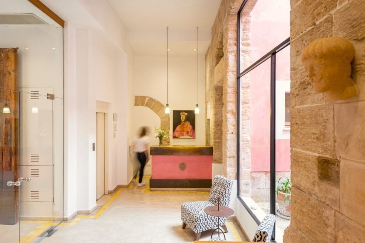 Contemporary hotel lobby with a pink reception desk, ancient stone walls, and a glass-enclosed area. A person walks past, and a patterned chair awaits.