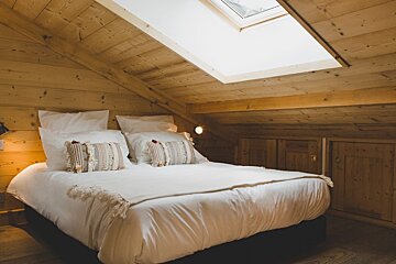 A cozy, wood-paneled attic bedroom features a large bed with white bedding, decorative pillows, a bright skylight, and built-in wooden cupboards.