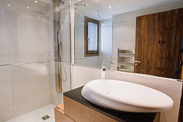 A modern bathroom features a white oval vessel sink on a wooden vanity, a glass shower, and a large mirror reflecting a window and wooden door.
