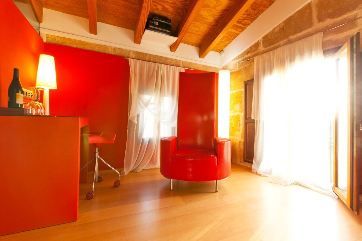 Bright room with bold red and orange decor, wooden floor, exposed beams, a tall red armchair, and sunny windows with white sheer curtains.