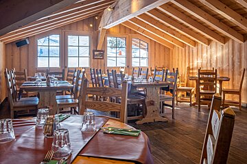 Cozy, rustic wooden restaurant with set tables, a sloped ceiling, and large windows offering views of a snowy outdoor landscape.