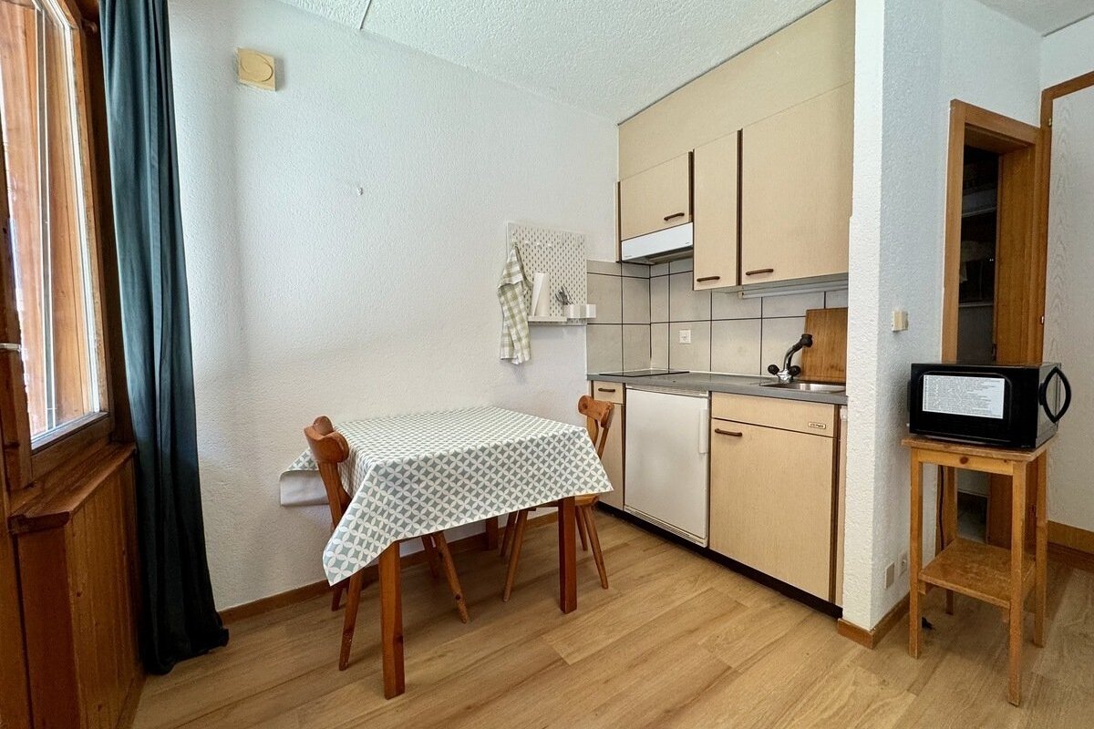 A small kitchenette and dining area with a wooden table, two chairs, fridge, sink, and microwave. Wooden floor and light cabinets.