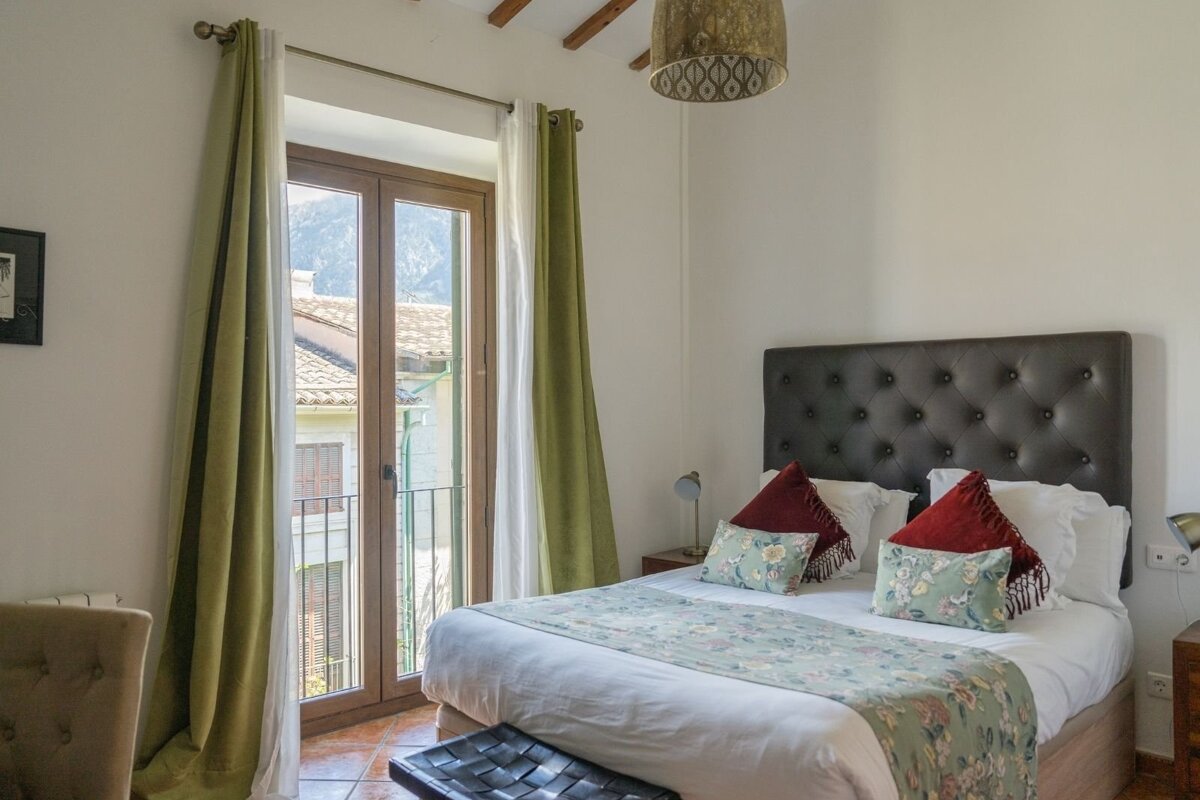 Bright bedroom with a dark tufted bed, floral runner, and a balcony door with green curtains revealing a mountain and village view.
