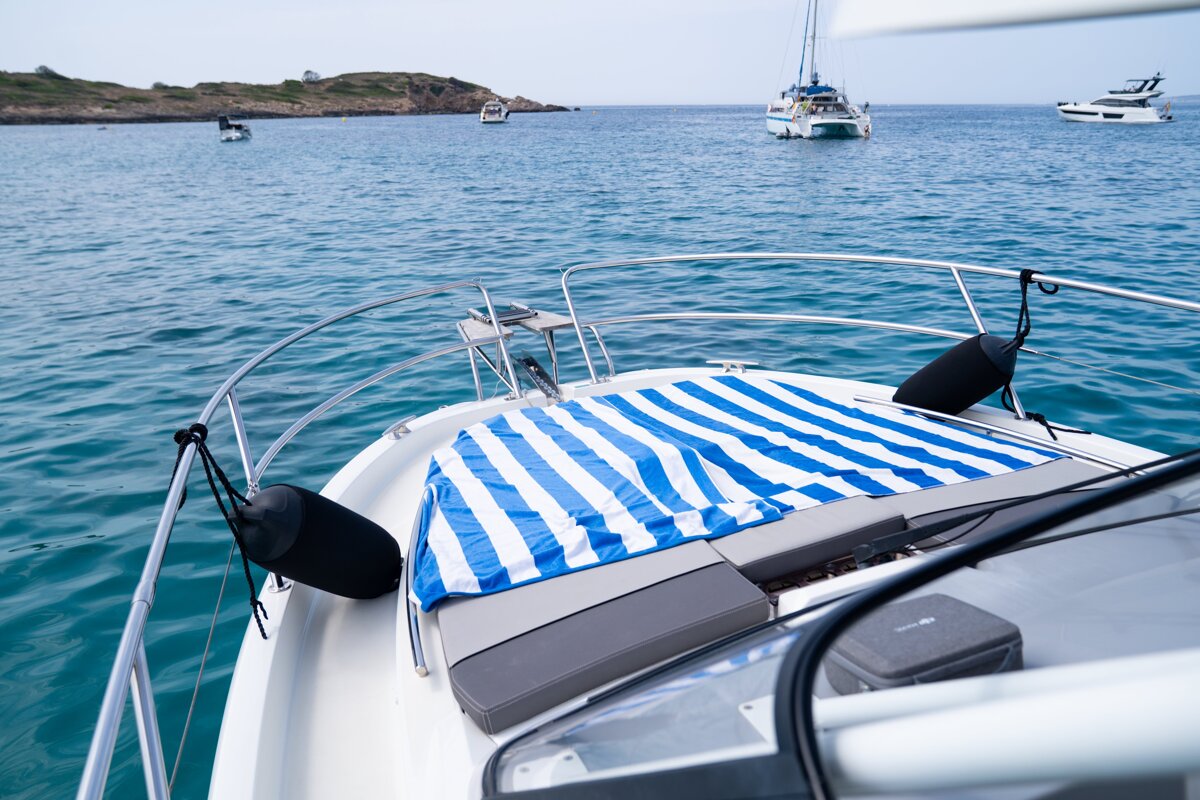 View from a boat's bow featuring a blue & white striped blanket. Blue ocean with other boats and an island in the distance.