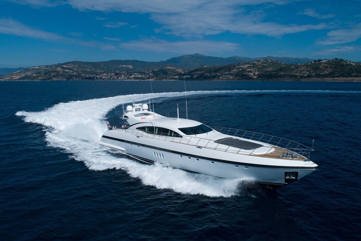 A sleek white yacht speeds across dark blue water, creating a foamy wake. A hilly coastline is visible in the background under a clear blue sky.