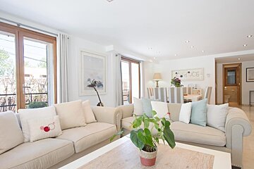 A bright, open living room and dining area featuring two beige sofas with pillows, a dining table with striped chairs, and large windows.