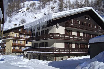 Snow-covered chalets with long icicles hanging from their roofs in a winter mountain village. Deep snow blankets the ground and distant hills with bare trees.