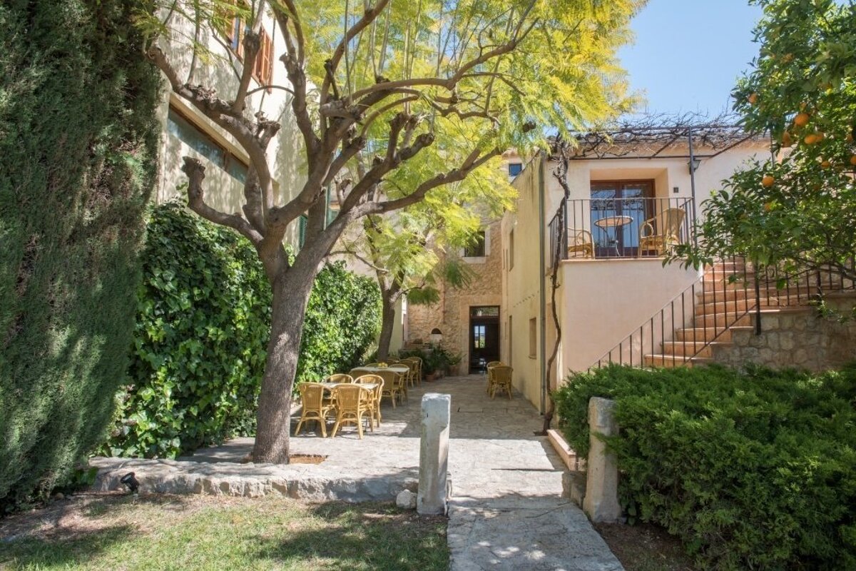 Sunny Mediterranean courtyard with light buildings, lush trees, and rattan outdoor dining areas. A balcony with seating is accessed via a stone staircase.