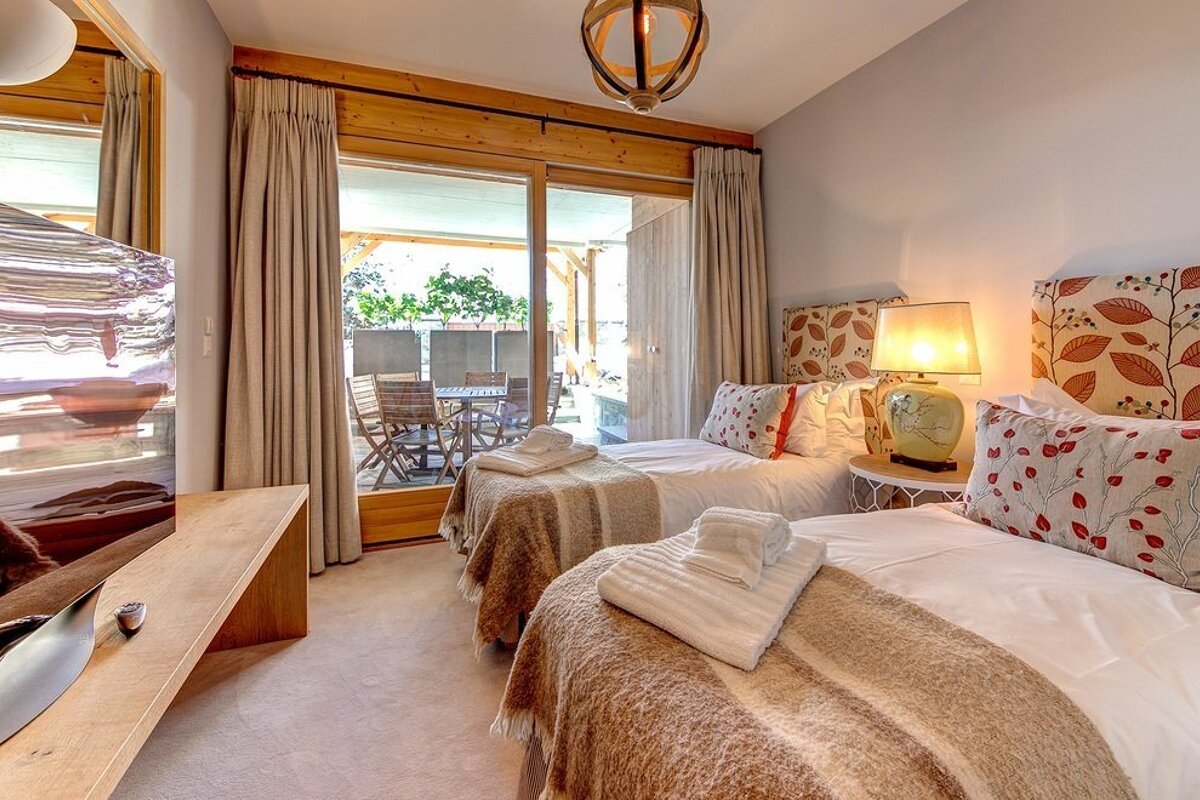 A cozy bedroom with two single beds, patterned headboards, a bedside lamp, and sliding glass doors opening to a patio with outdoor furniture.