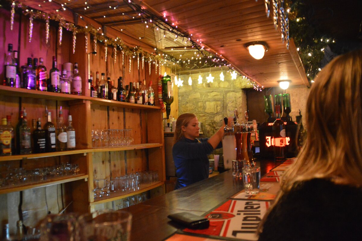 A bartender serves drinks at a festive wooden bar adorned with Christmas lights and icicles, with shelves of liquor bottles and glasses visible.