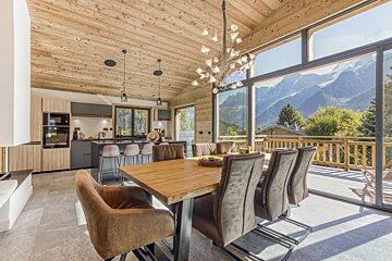 A modern, open-plan chalet interior with a dining area, kitchen, and large windows revealing breathtaking panoramic mountain views.