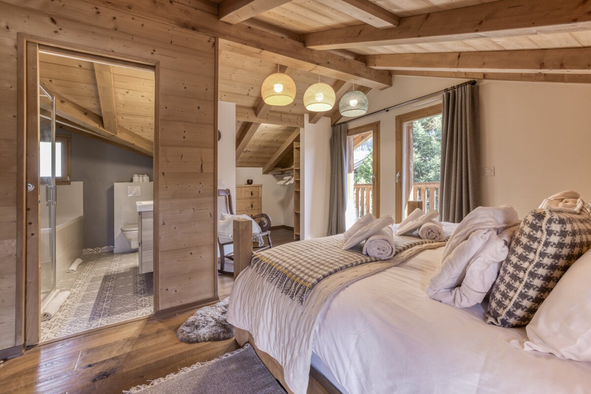 A cozy, wood-paneled bedroom with exposed beams features an open en-suite bathroom, a plush bed with towels, and double doors to a balcony.