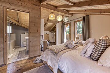 A cozy, wood-paneled bedroom with exposed beams features an open en-suite bathroom, a plush bed with towels, and double doors to a balcony.