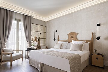 A serene, elegant bedroom in neutral tones with a wooden bed, large window with drapes, a comfy armchair, and a decorative mirrored wall panel.