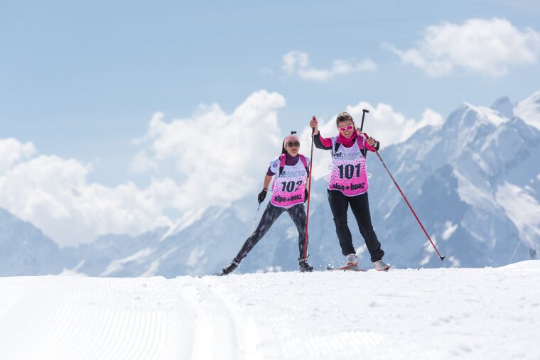 Two women biathlon skiing in pink bibs (101 & 102) on a sunny, snowy mountain slope with majestic peaks and clouds in the background.