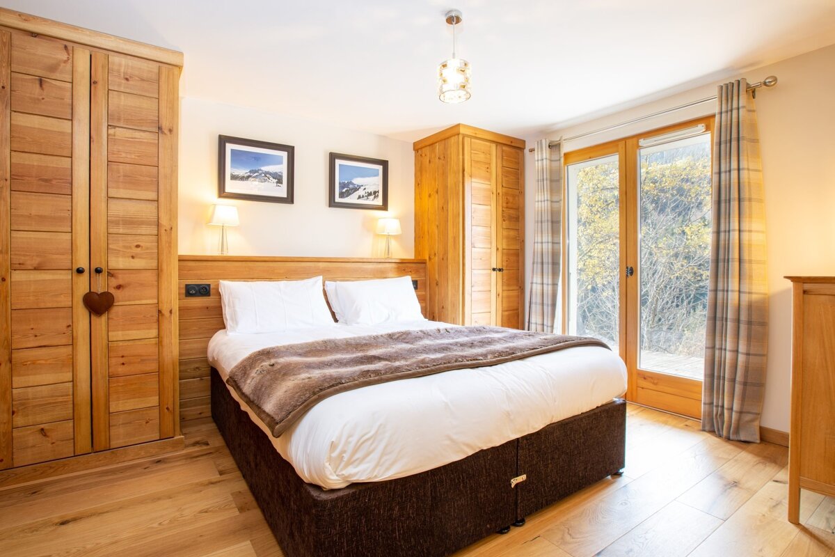 A bright, cozy bedroom featuring a wooden bed, wardrobes, and a large glass door looking out onto trees. Mountain artwork and warm lighting create a natural ambiance.