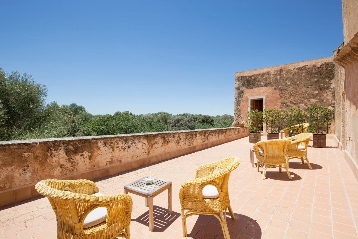 Sunny outdoor terrace with wicker chairs on terracotta tiles, a rustic building, and a panoramic view of green trees under a clear blue sky.