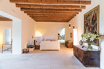 A bright, rustic living room features exposed wooden beams, white walls, a white sofa, and a large painting above dark wooden chests.