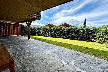 A stone patio under a wooden overhang looks out over a green lawn and hedge toward mountain cabin roofs under a cloudy sky.
