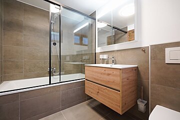 A clean, modern bathroom with brown tiles, a bathtub/shower combo, a wooden floating vanity with sink, and an illuminated mirror cabinet.