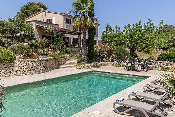 A sunny Mediterranean villa features a sparkling pool, surrounded by stone walls, lush gardens, and palm trees, with lounge chairs and an outdoor dining area.