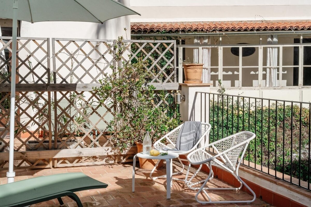 Cozy outdoor patio with white chairs, table, and umbrella, surrounded by lush plants and a lattice fence, with a building visible beyond.