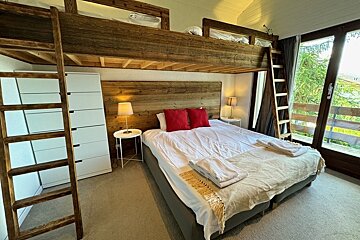 A rustic-modern bedroom features wooden bunk beds built over a main bed, red pillows, and bedside tables with lamps, beside a large window.