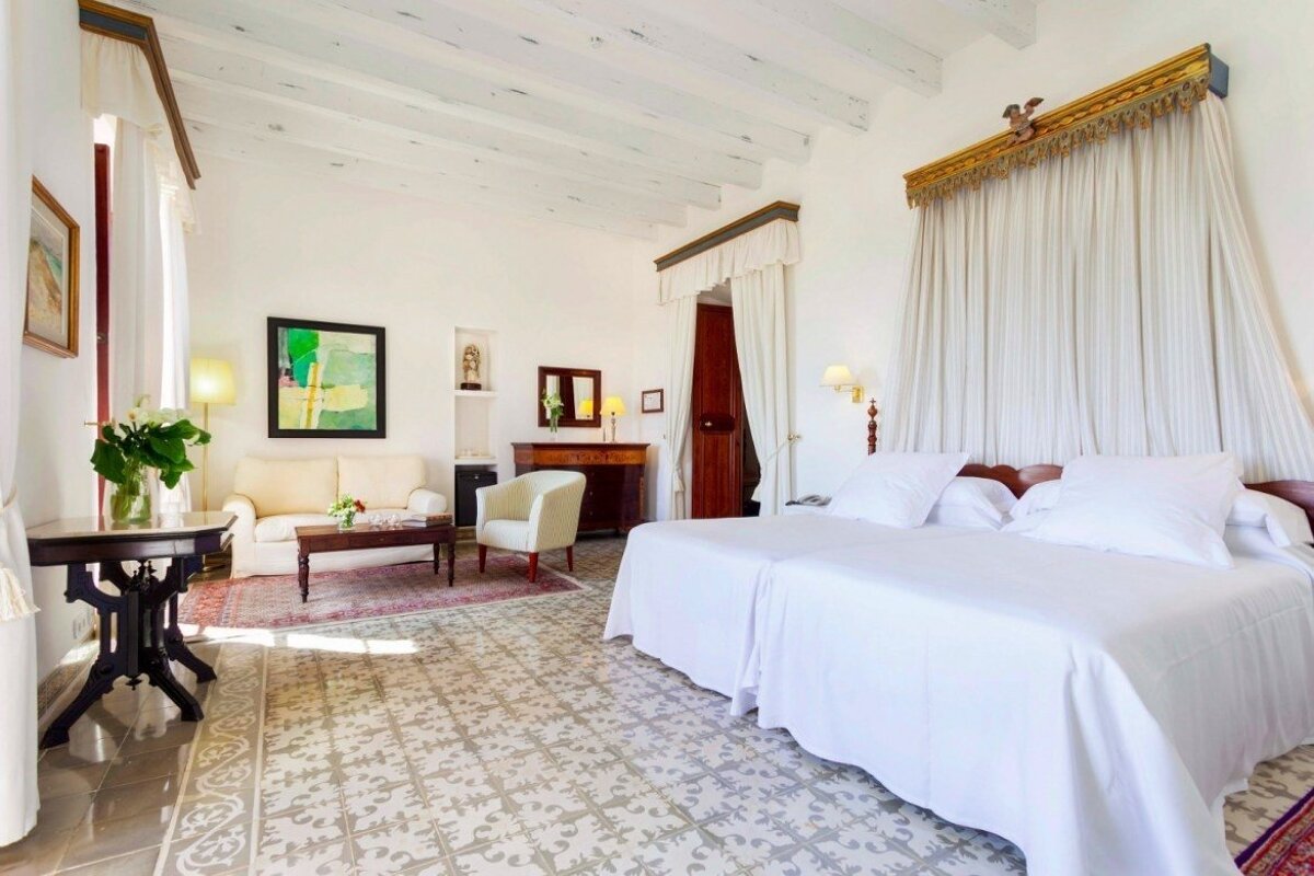 An elegant hotel room featuring twin beds under a draped canopy, a separate sitting area with a sofa, patterned floor tiles, and a beamed ceiling.