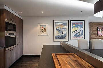 Modern kitchen with wood cabinets, integrated microwave/oven, dark counter with cutting board. Vintage ski posters on wall, dining area nearby.