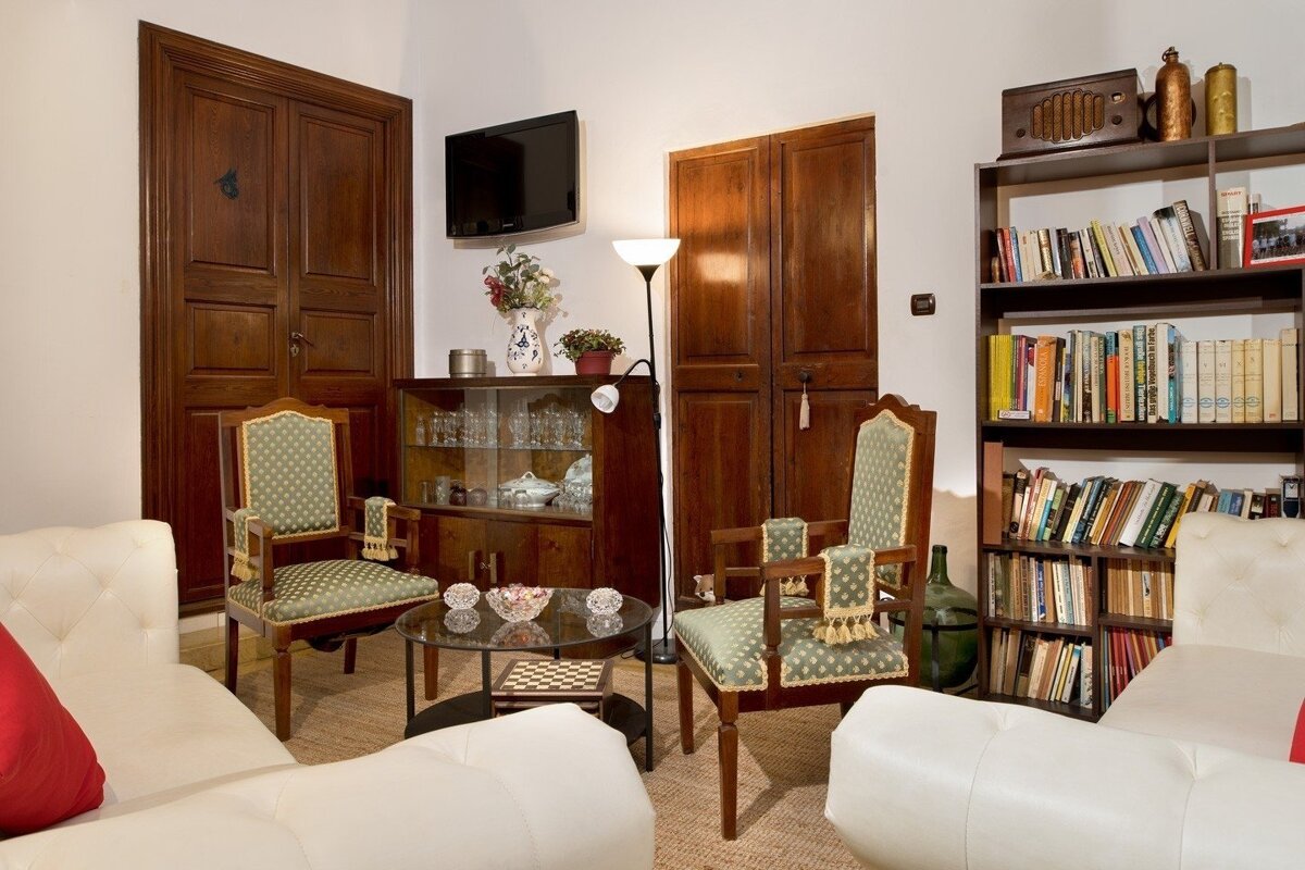 A traditional living room with white sofas, green patterned armchairs, dark wood furniture including a bookshelf, a glass coffee table, and a wall-mounted TV.