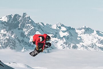 A snowboarder in a red jacket jumps mid-air against a spectacular backdrop of snow-capped mountains.