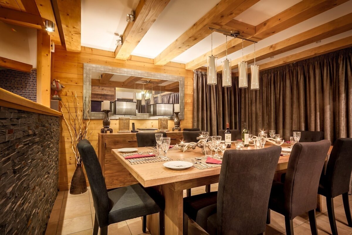 A warm, inviting wooden dining room with exposed beams, a set table, chic chandelier, and large mirror. Stone accents add rustic charm.