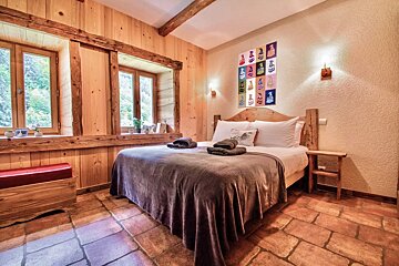 A rustic wooden bedroom featuring a large bed, terracotta tiled floor, and windows overlooking greenery. Pop-art decor adds a modern touch.