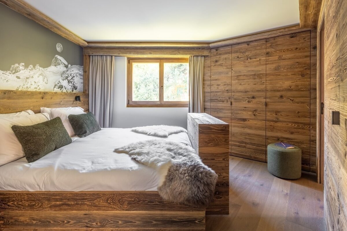 A cozy, rustic alpine bedroom with a wooden bed, white linens, and fur throws. A mountain mural and large window complement the wooden wardrobe.