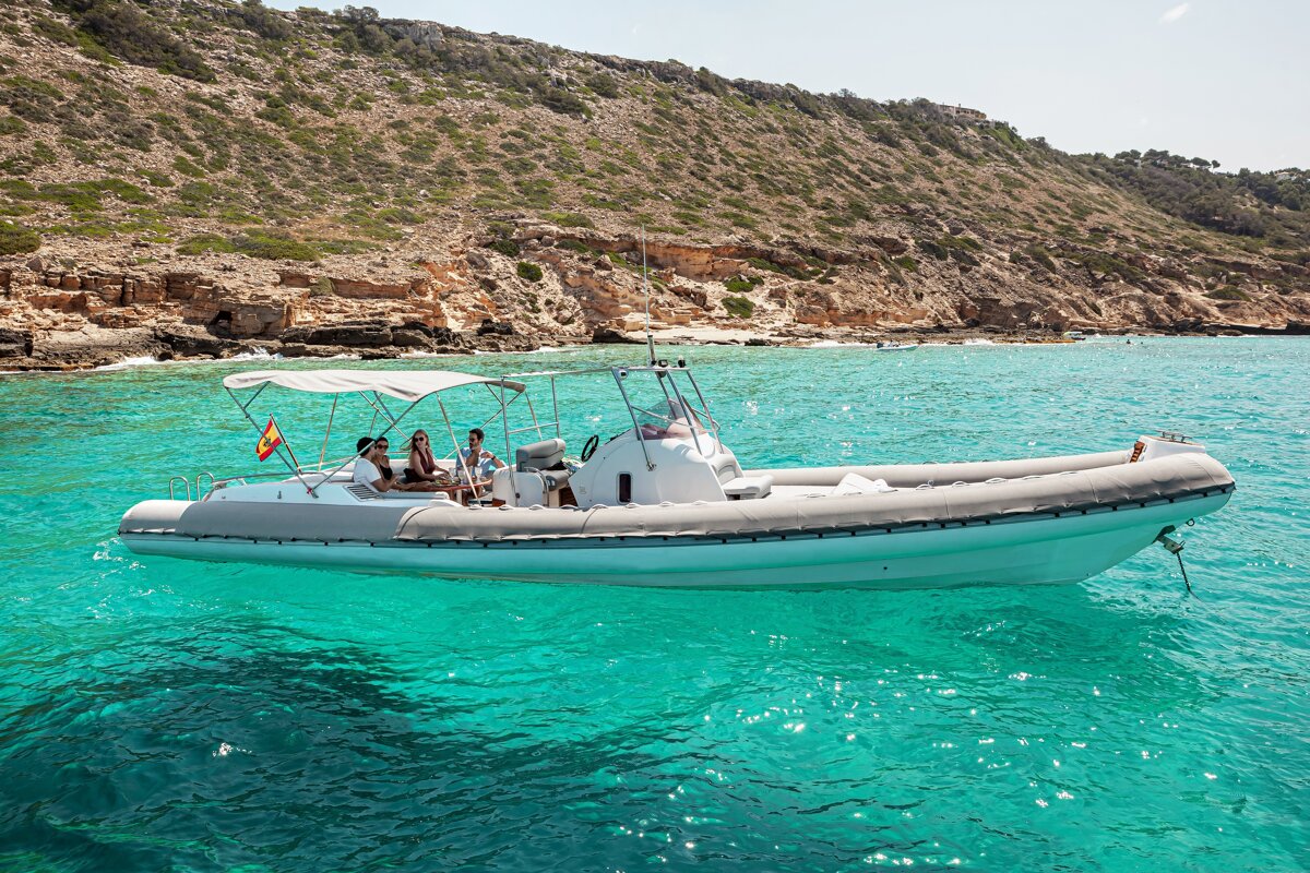People relax on a rigid inflatable boat floating in crystal-clear turquoise water near a rocky coastline.