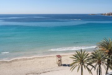 A tranquil, aerial view of a sandy beach with clear turquoise water, palm trees, and a lifeguard tower, with a distant city and boat.