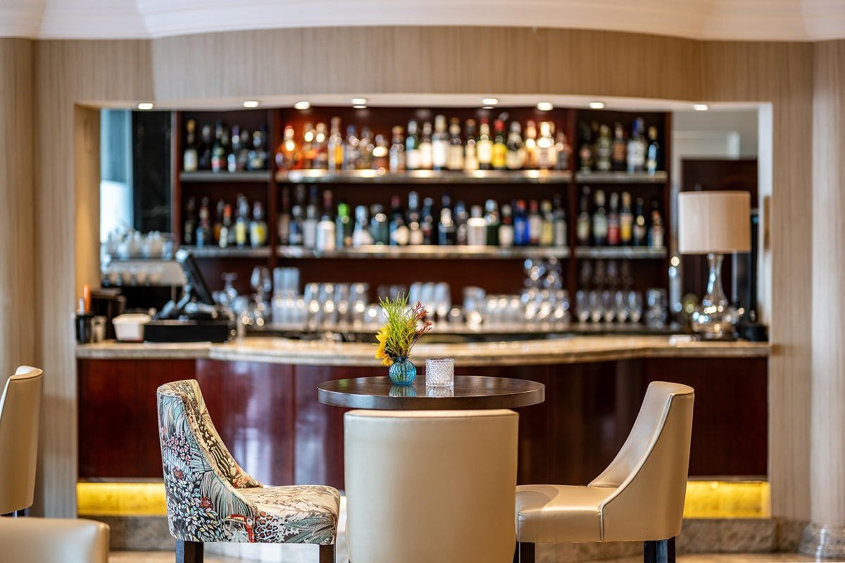 A stylish bar area with shelves of liquor bottles, glasses, and elegant seating in the foreground, including a table with a small flower vase.