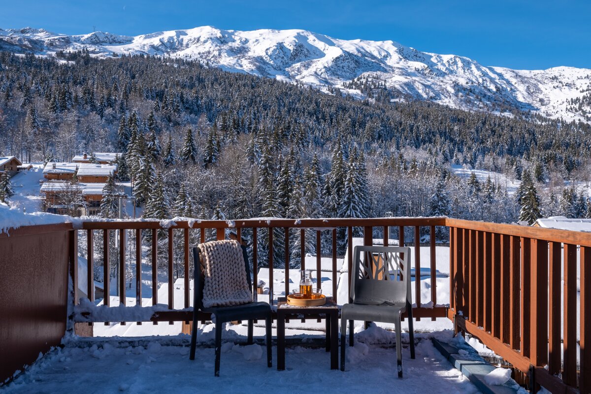 A snowy balcony with two chairs overlooking a dense winter forest and majestic snow-covered mountains under a bright blue sky.