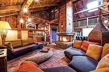 A spacious, rustic chalet living room with exposed wooden beams, a grand fireplace, and colorful, plush seating, offering warmth and comfort.