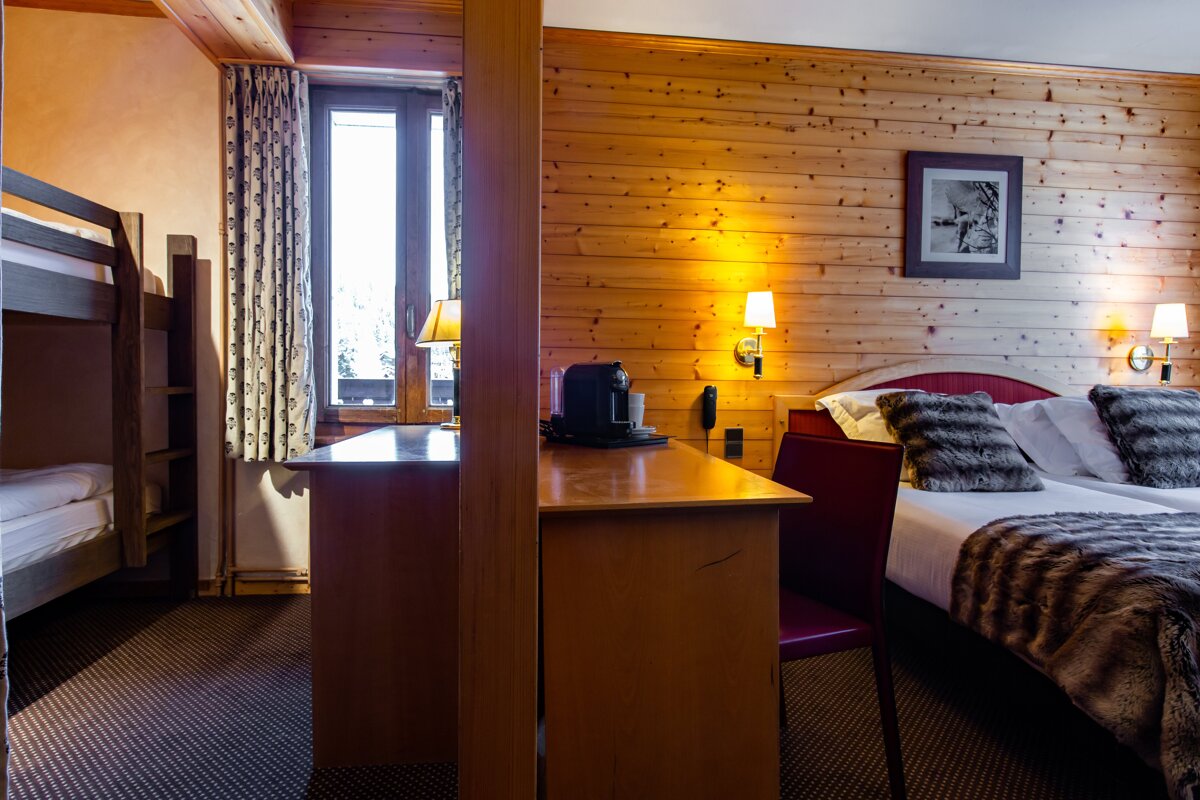 A warm, wood-paneled hotel room with a double bed, bunk beds, a desk, and a coffee machine. A bright window is visible.