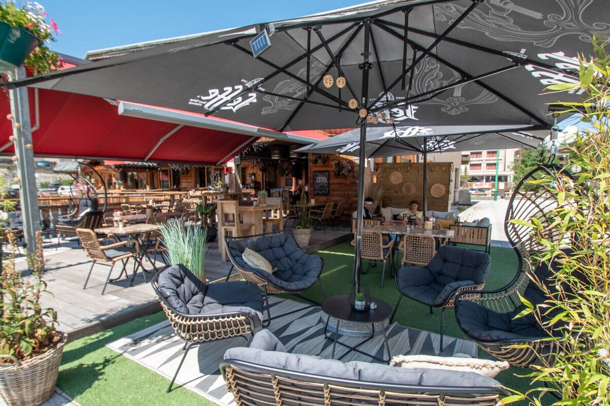 A sunny outdoor cafe patio featuring black umbrellas, red awnings, wicker and wooden seating areas, lush plants, and a mix of artificial turf and wooden decking.